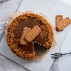 Pyszny sernik z polewą karmelową i pokruszonymi ciastkami Lotus. Obok leżą dwa całe ciastka Lotus i widelec.