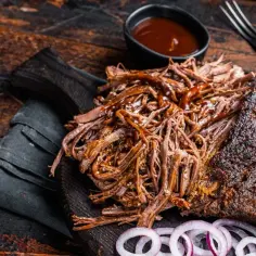 Soczysta, szarpana wołowina z sosem BBQ na drewnianej desce, obok plasterki cebuli i miseczka sosu. Idealna na grilla.