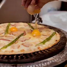 Ręka wylewa jajko na pizzę z bekonem i szparagami. Pyszna pizza z jajkiem w trakcie przygotowania.