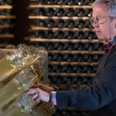 Starszy mężczyzna w okularach przegląda butelki z winem musującym. W piwnicy pełnej regałów z winem, czeka na degustację cava.