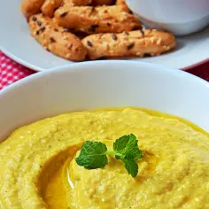 Gładki, żółty hummus z fava fasoli, ozdobiony listkiem mięty, podany z chrupiącymi paluszkami na talerzu.