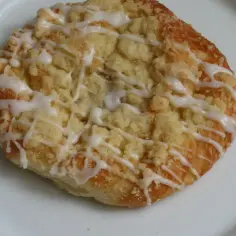 Szneka z glancem, puszyste ciasto drożdżowe z kruszonką i lukrem, wygląda apetycznie na białym talerzu.
