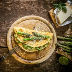 Pyszny omlet ze szparagami, posypany parmezanem, na drewnianej desce. Obok pęczek świeżych szparagów i kawałek sera.