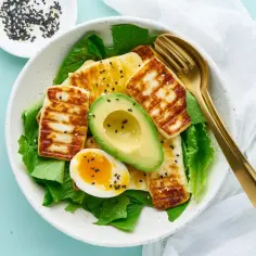 Pyszne keto śniadanie: grillowany ser halloumi, awokado i jajko na miękko na sałacie, posypane sezamem. Złote sztućce dodają elegancji.