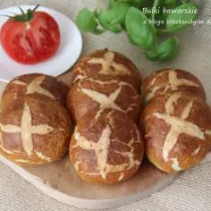 Acht bułek bawarskich z charakterystycznym nacięciem, ułożonych na drewnianej desce. Obok pomidor i listki bazylii.