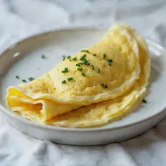 Delikatny omlet francuski, złożony na pół, posypany szczypiorkiem, na białym talerzu.